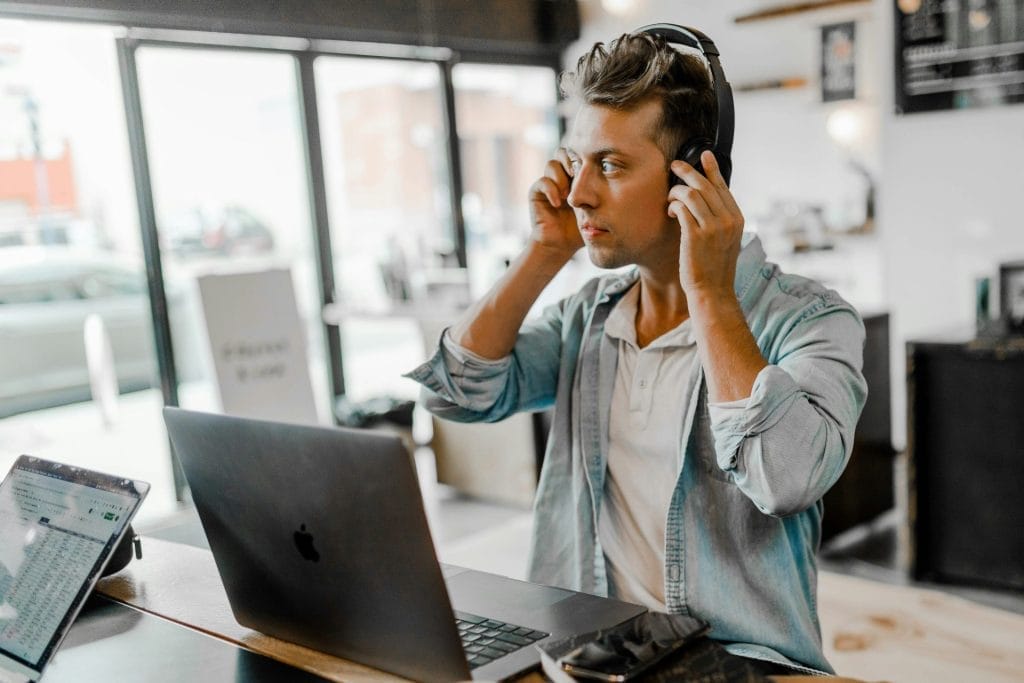 A man is sitting at a desk in front of an open laptop, wearing a light denim shirt. He is adjusting his headphones with both hands, ready for a virtual meeting on maximizing revenue as an Airbnb host. The modern, well-lit workspace includes large windows, a tablet, and a smartphone on the desk.