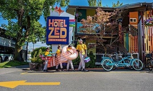 Four people in yellow shirts carry large inflatable pool toys in front of a colorful building with a sign reading "Hotel Zed Victoria." With several bicycles parked nearby and a bus visible on the left, the vibrant scene hints at the property's revenue potential through its unique and inviting atmosphere.