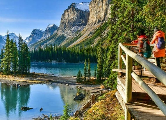 Tourists stand on a wooden deck overlooking a scenic view of a turquoise lake, surrounded by dense pine forests and rugged mountains. The clear, bright sky enhances this breathtaking natural beauty, offering moments that even a hotel revenue management expert would find priceless.