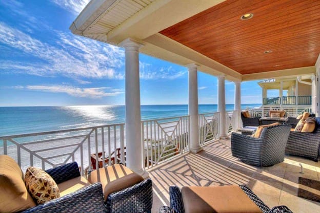 Spacious beachside balcony with wicker lounge chairs and cushions, white columns, and a wooden ceiling—overlooking a bright blue ocean and sunny sky, ideal for listings from top Vacation Rental Management Companies in Canada.